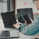 Person working across multiple laptops, a monitor, and a phone at once, representing how NTP keeps every connected device synchronized to the same time