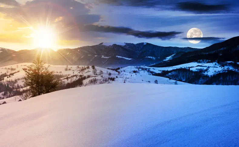 Sun and full moon visible together in the sky over a snow-covered mountain landscape, illustrating the seasonal shift in daylight through the year