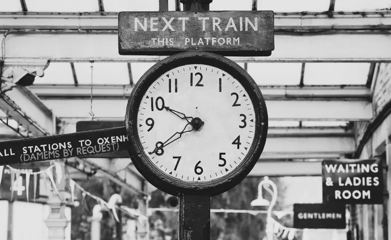 Vintage railway station clock beneath a "Next Train This Platform" sign, symbolizing how railroads imposed standardized time on a world of clashing local clocks
