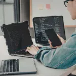 Person working across multiple laptops, a monitor, and a phone at once, representing how NTP keeps every connected device synchronized to the same time