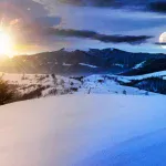 Sun and full moon visible together in the sky over a snow-covered mountain landscape, illustrating the seasonal shift in daylight through the year