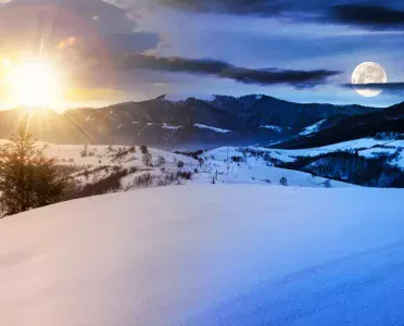 Sun and full moon visible together in the sky over a snow-covered mountain landscape, illustrating the seasonal shift in daylight through the year