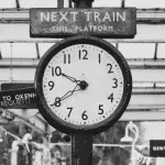 Vintage railway station clock beneath a "Next Train This Platform" sign, symbolizing how railroads imposed standardized time on a world of clashing local clocks