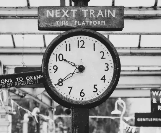 Vintage railway station clock beneath a "Next Train This Platform" sign, symbolizing how railroads imposed standardized time on a world of clashing local clocks